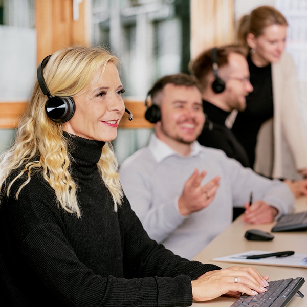 Eine Blonde Frau aus dem Customer Management Team führt  offenbar ein Kundengespräch mit Headset. Im Hintergrund sind drei weitere Personen aktiv in Gesprächen involviert